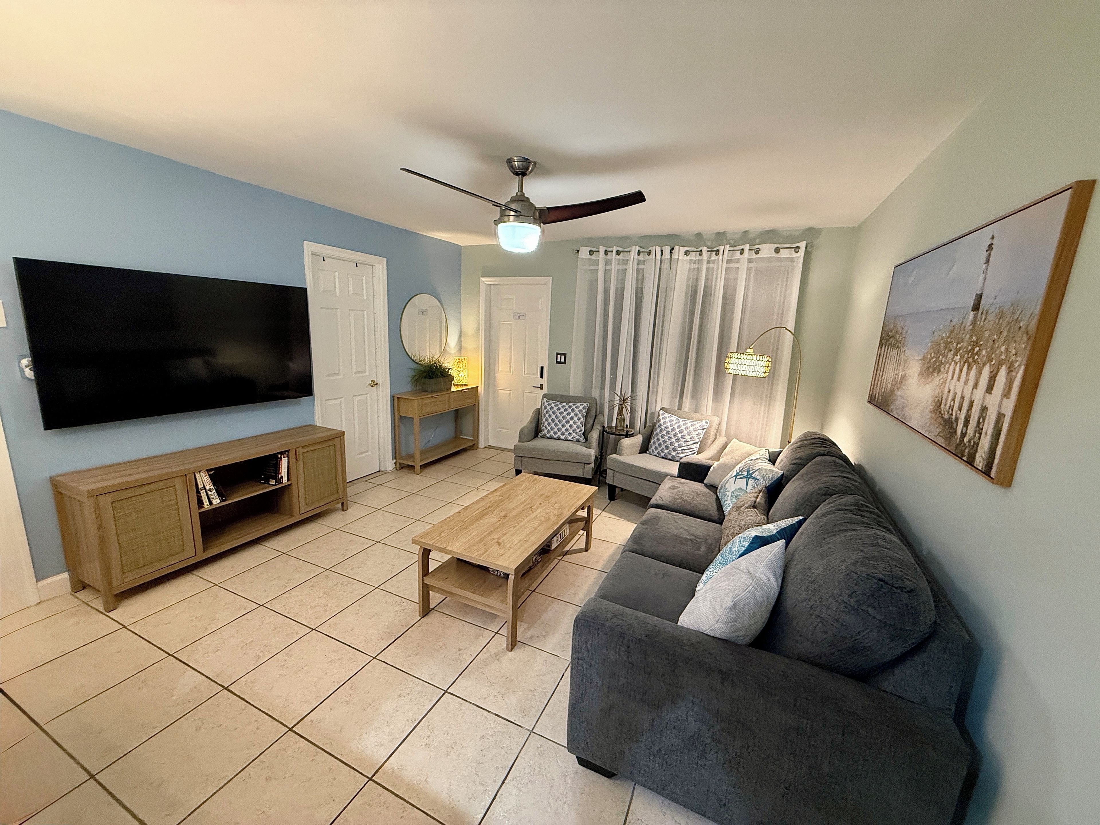 A living room with a flat-screen TV, a wooden cabinet, a grey sofa, a coffee table, and a ceiling fan.