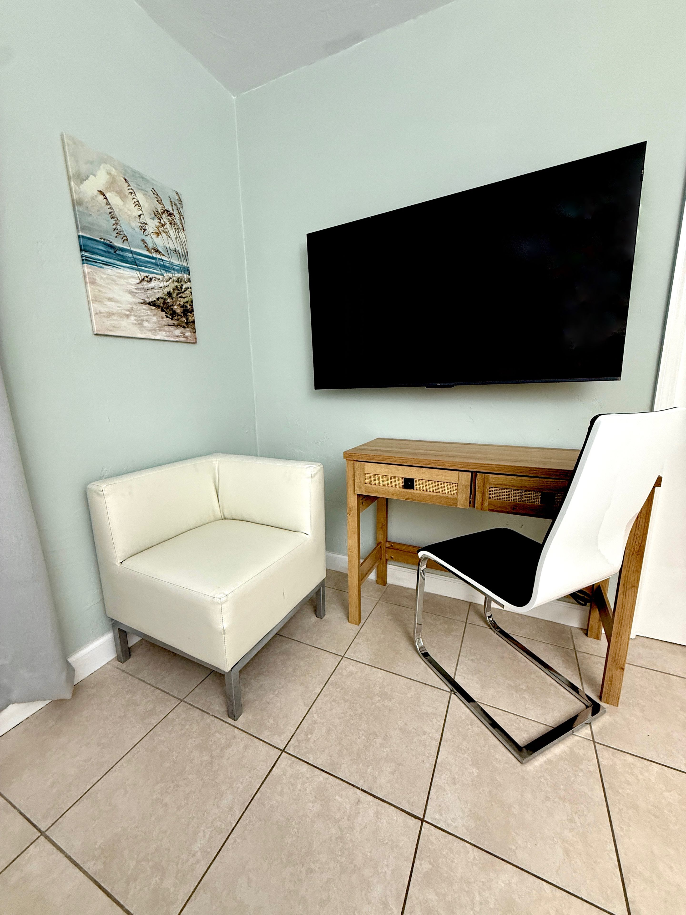 A room with a white leather sofa, a wooden desk with drawers, a white chair, a flat-screen TV mounted on the wall, and a painting of a beach scene.