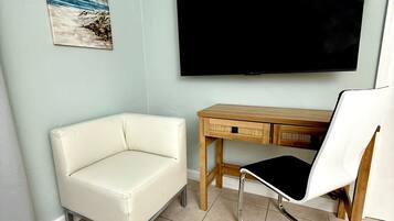 A room with a white leather sofa, a wooden desk with drawers, a white chair, a flat-screen TV mounted on the wall, and a painting of a beach scene.
