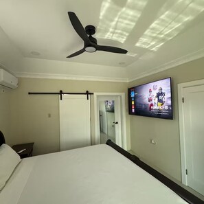A hotel room with a bed, a ceiling fan, a mounted TV displaying a sports event, and a sliding door. - Trenz De Luxe (May Pen)