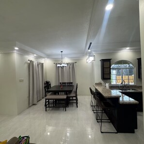 A modern kitchen with a dining area, a large window, and a built-in island with bar stools. - Trenz De Luxe (May Pen)