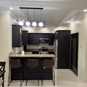 A modern kitchen with dark cabinetry, a granite countertop, a stainless steel refrigerator, and a bar area with stools. - Trenz De Luxe (May Pen)