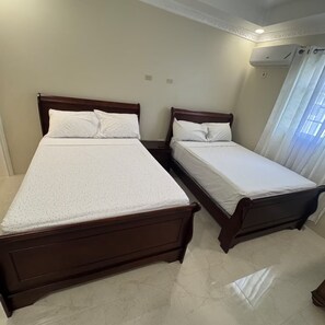 A bedroom with two wooden beds, a white ceiling, and a window with curtains. - Trenz De Luxe (May Pen)