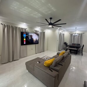 A modern living room with a flat-screen TV, a ceiling fan, a dining area, and a sofa with cushions. - Trenz De Luxe (May Pen)