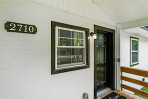 Una casa blanca con puerta y ventana negras, un cartel con el número '2710' sobre la puerta y un banco de madera afuera.