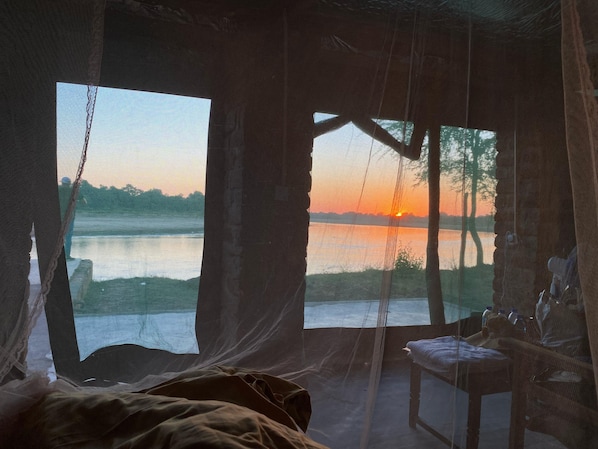 A room with a bed, a chair, and a view of a sunset over a body of water. - Mikango safaris (South Luangwa National Park)