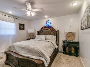 A bedroom with a large bed, a ceiling fan, a stained glass window, and a small side table. - Spacious 7-bedroom home with heated pool and Jaccuzzi (Oklahoma City)