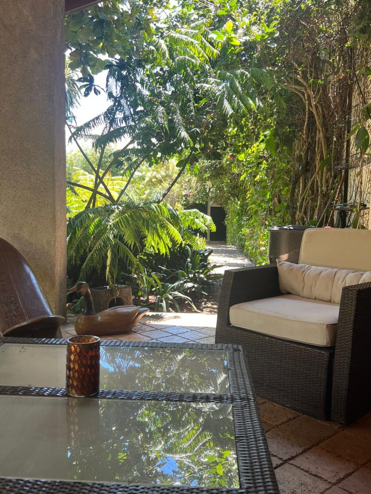 A glass-top table with a decorative bowl and a patterned glass container, a wicker sofa with cushions, and a view of lush greenery and a pathway leading to a door.