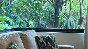 Una habitación con un sofá color crema, una mesa de centro de vidrio y vistas a un follaje verde exuberante a través de un ventanal grande.
