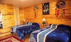 A room with wooden walls, two beds with blue bedding, a nightstand, a lamp, and wall decorations including dreamcatchers and framed pictures. - Hotel Cabanas Taramuri (Bocoyna)