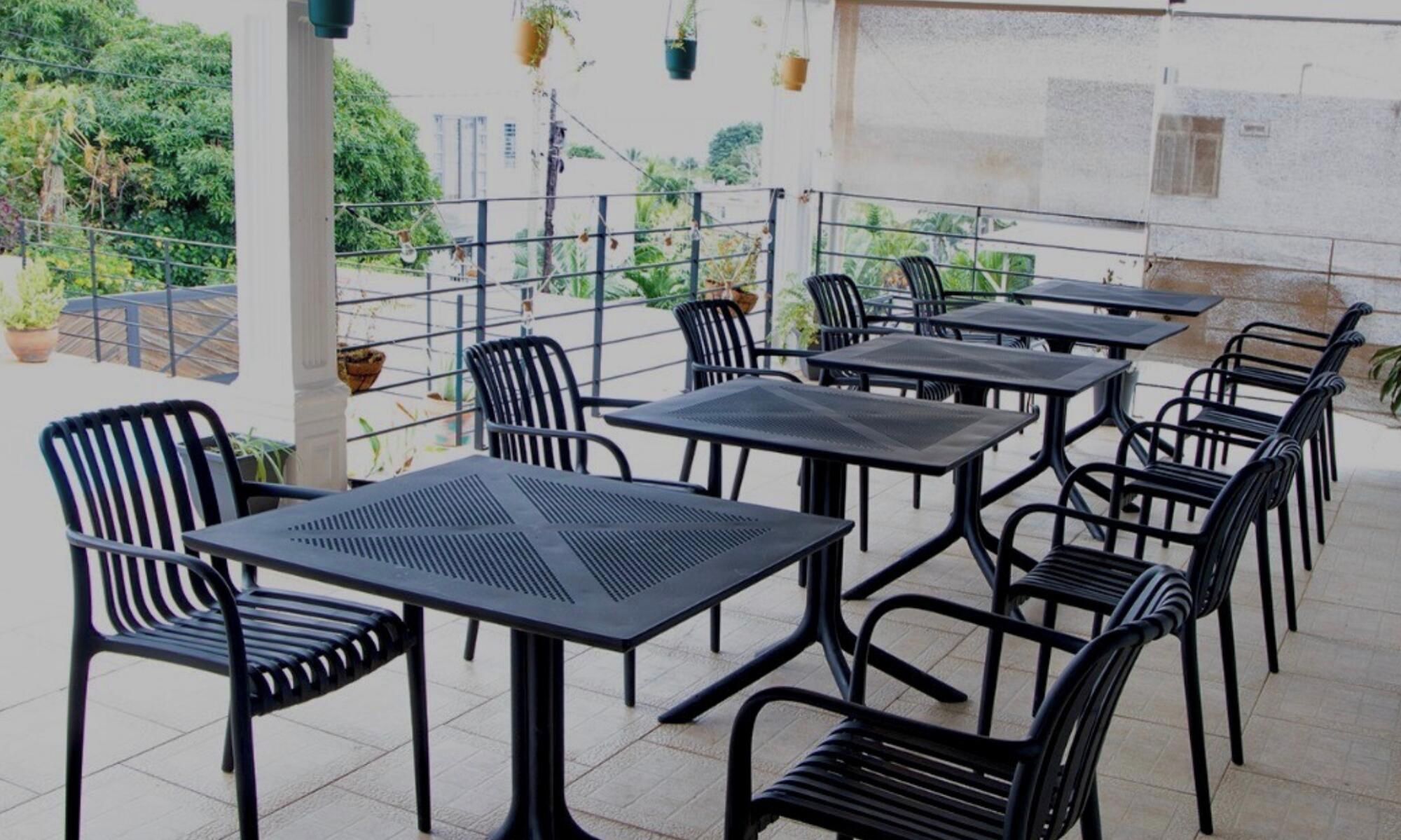 On peut apercevoir une terrasse extérieure avec plusieurs tables et chaises, entourée d’une rambarde en métal, et des plantes en pot en arrière-plan.