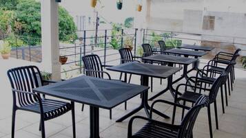 On peut apercevoir une terrasse extérieure avec plusieurs tables et chaises, entourée d’une rambarde en métal, et des plantes en pot en arrière-plan.