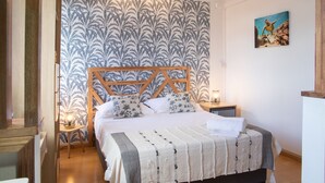 Un dormitorio con una cama, cabecera de madera, papel tapiz estampado, mesita de noche, lámpara y un cuadro en la pared.