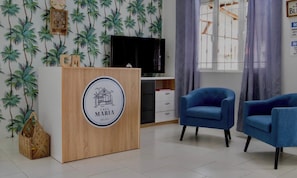 A room with a wooden reception desk, two blue armchairs, a television, and a window with curtains.