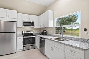 A modern kitchen with stainless steel appliances, white cabinetry, a granite countertop, and a window with a view of the outdoors. - 2 Houses Shark Tooth Acres - Manasota Key Sleep 22 (Englewood)