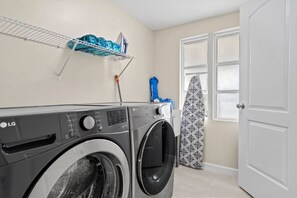 A laundry area with a front-loading washer and dryer, a shelf with folded clothes, and an ironing board. - 2 Houses Shark Tooth Acres - Manasota Key Sleep 22 (Englewood)