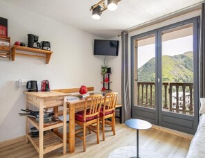 A cozy room with a wooden table, chairs, a small round table, a TV mounted on the wall, and a balcony with a view of hills. - Alpine Ski Apartment in Tignes for 4 (Saint-Sorlin-d'Arves)