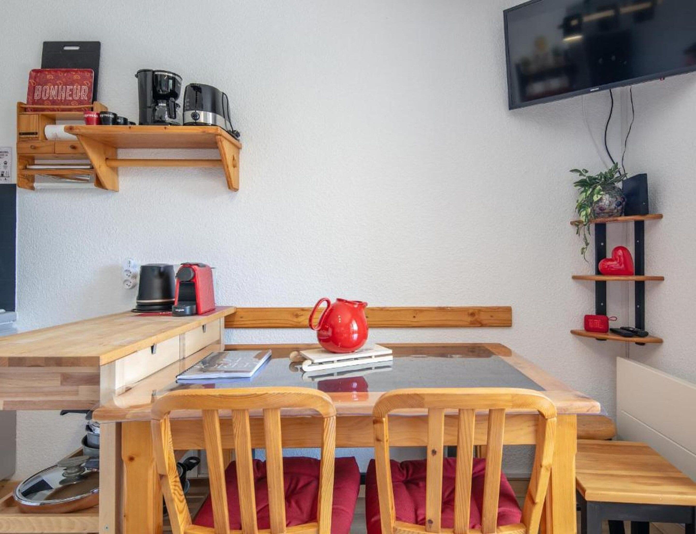 A compact living space with a wooden table and chairs, a coffee machine, a red kettle, and a flat-screen TV mounted on the wall.