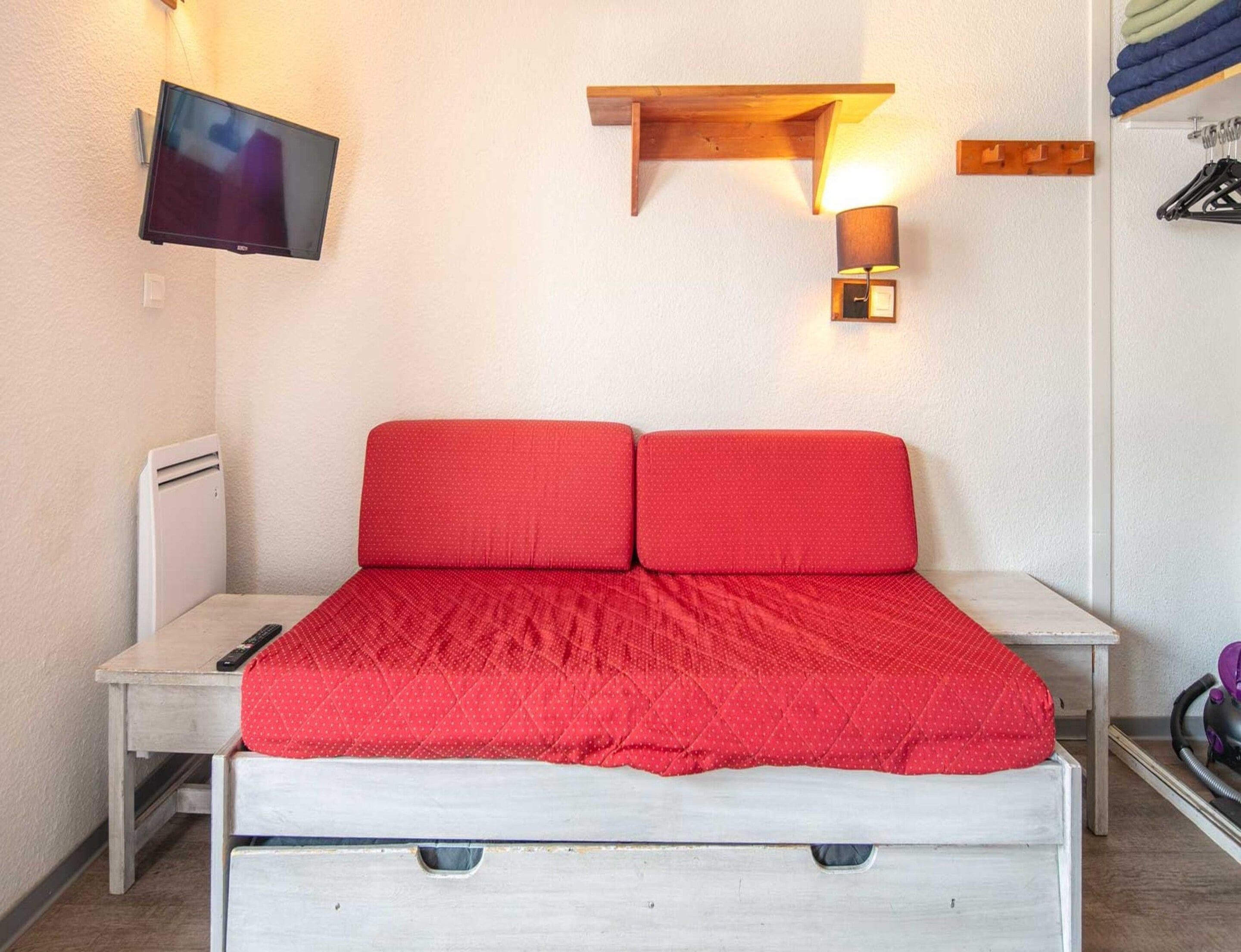 A room with a bed, a TV mounted on the wall, a shelf with a lamp, and a heater.
