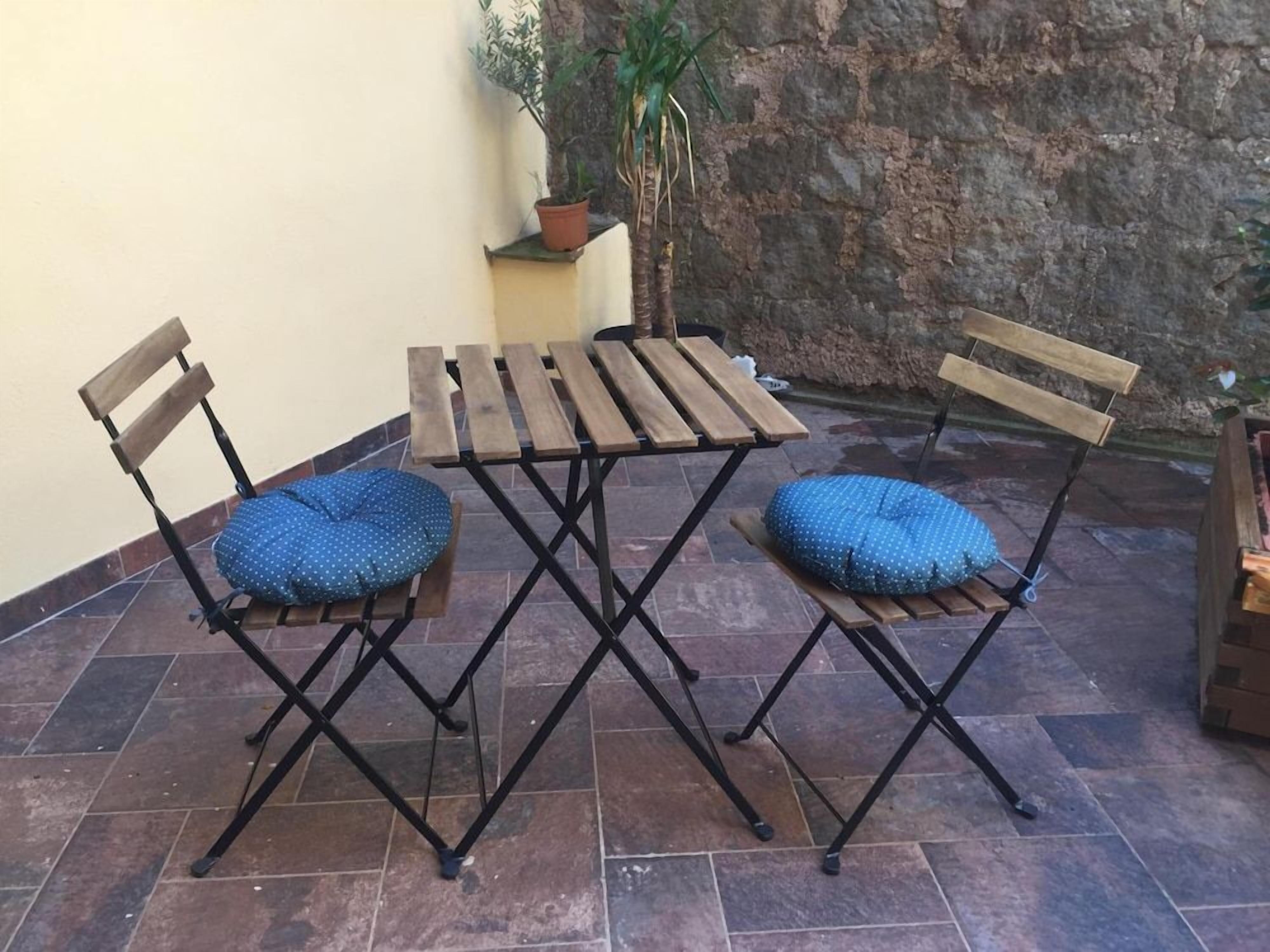 Un patio con una mesa de madera y dos sillas, una planta pequeña en maceta y un muro de piedra.