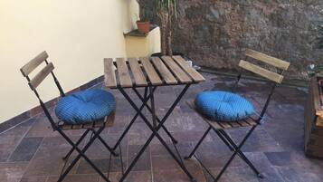 Um pátio com mesa de madeira e duas cadeiras, uma pequena planta em vaso e uma parede de pedra.