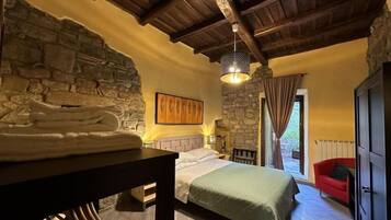 Quarto com parede de pedra, cama com manta verde, mesa de madeira com toalhas, cadeira vermelha e luminária pendente.