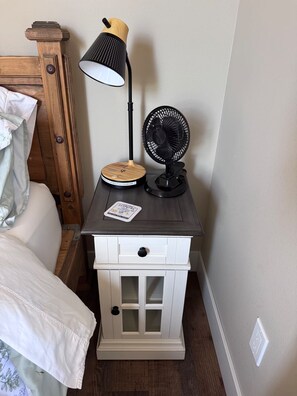 Una mesita de noche con lámpara, ventilador y cargador inalámbrico.