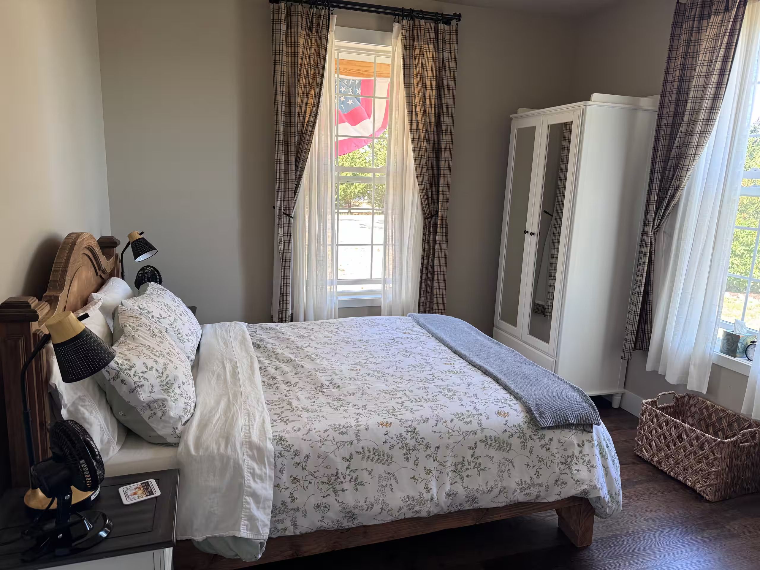 Un dormitorio con cama, armario, ventana con cortinas y mesita de noche con lámpara y ventilador.
