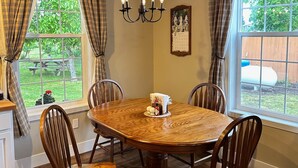 Uma sala de jantar com mesa de madeira, seis cadeiras, um lustre e grandes janelas com cortinas xadrez.