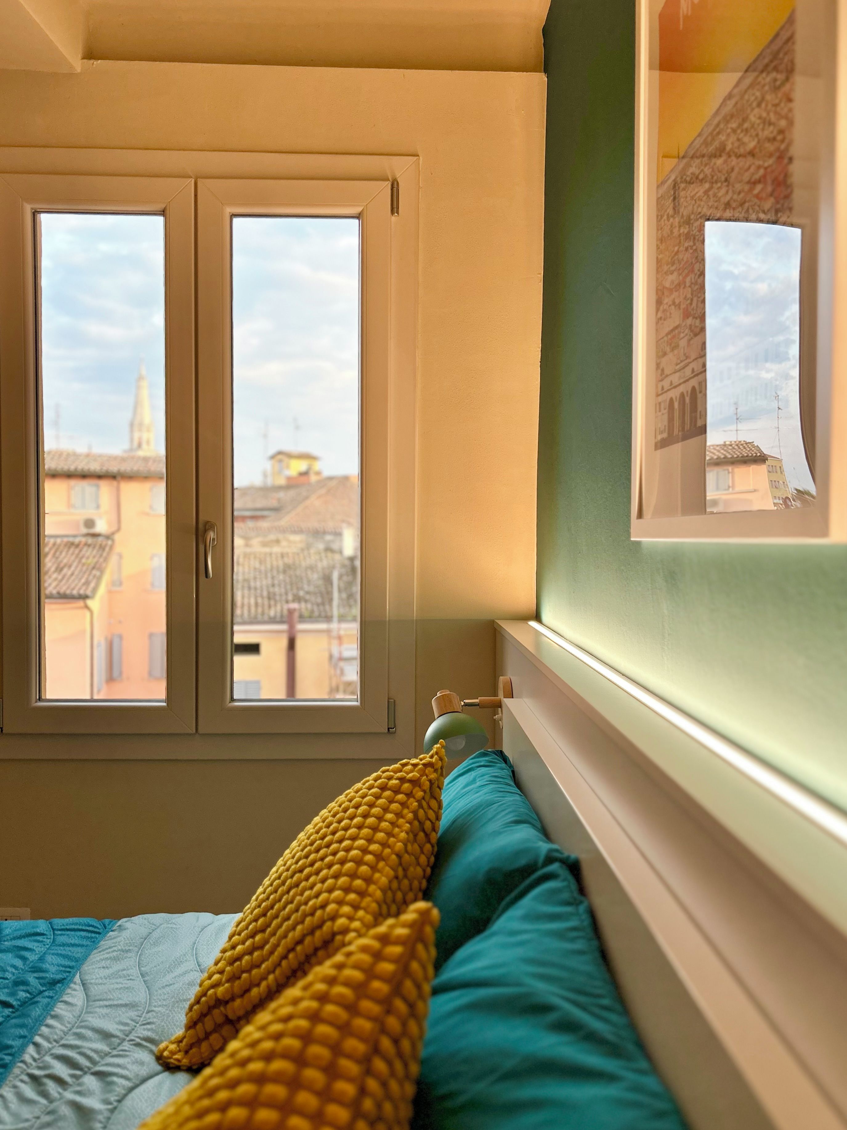 A bedroom with a bed, two pillows, a window, and a view of buildings outside.