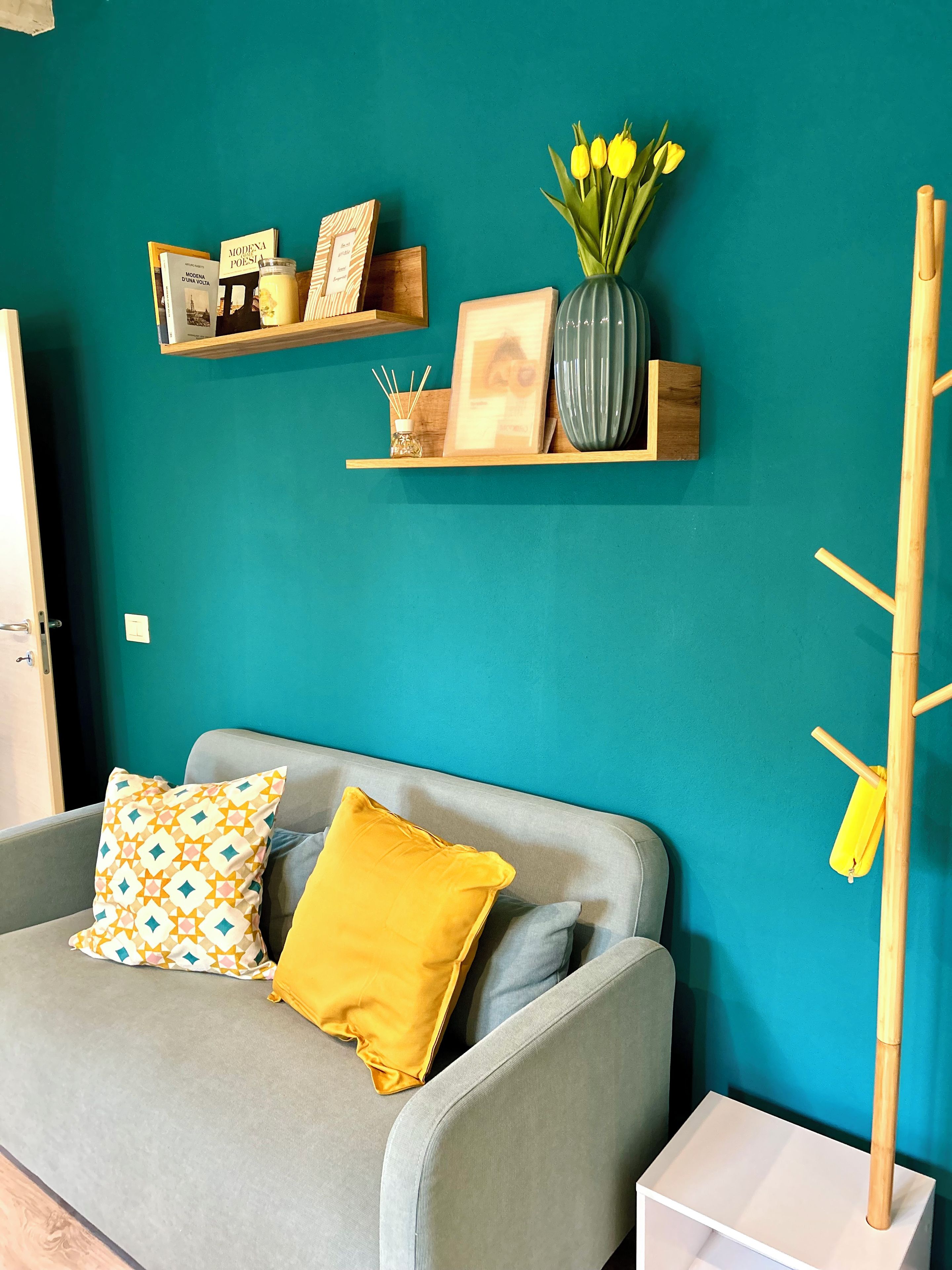 A teal-walled room with a grey sofa, patterned cushions, a vase with yellow flowers, and wooden shelves with books and decorative items.