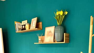 A teal-walled room with a grey sofa, patterned cushions, a vase with yellow flowers, and wooden shelves with books and decorative items.