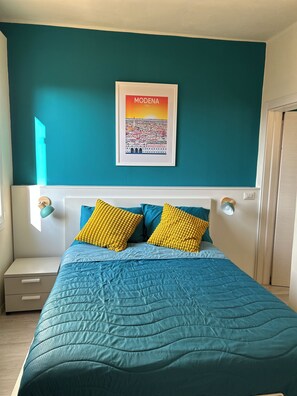 Une chambre avec un mur de couleur turquoise, une affiche encadrée représentant un paysage urbain, un lit avec une couette et des oreillers turquoise et jaunes, une table de chevet et une commode.