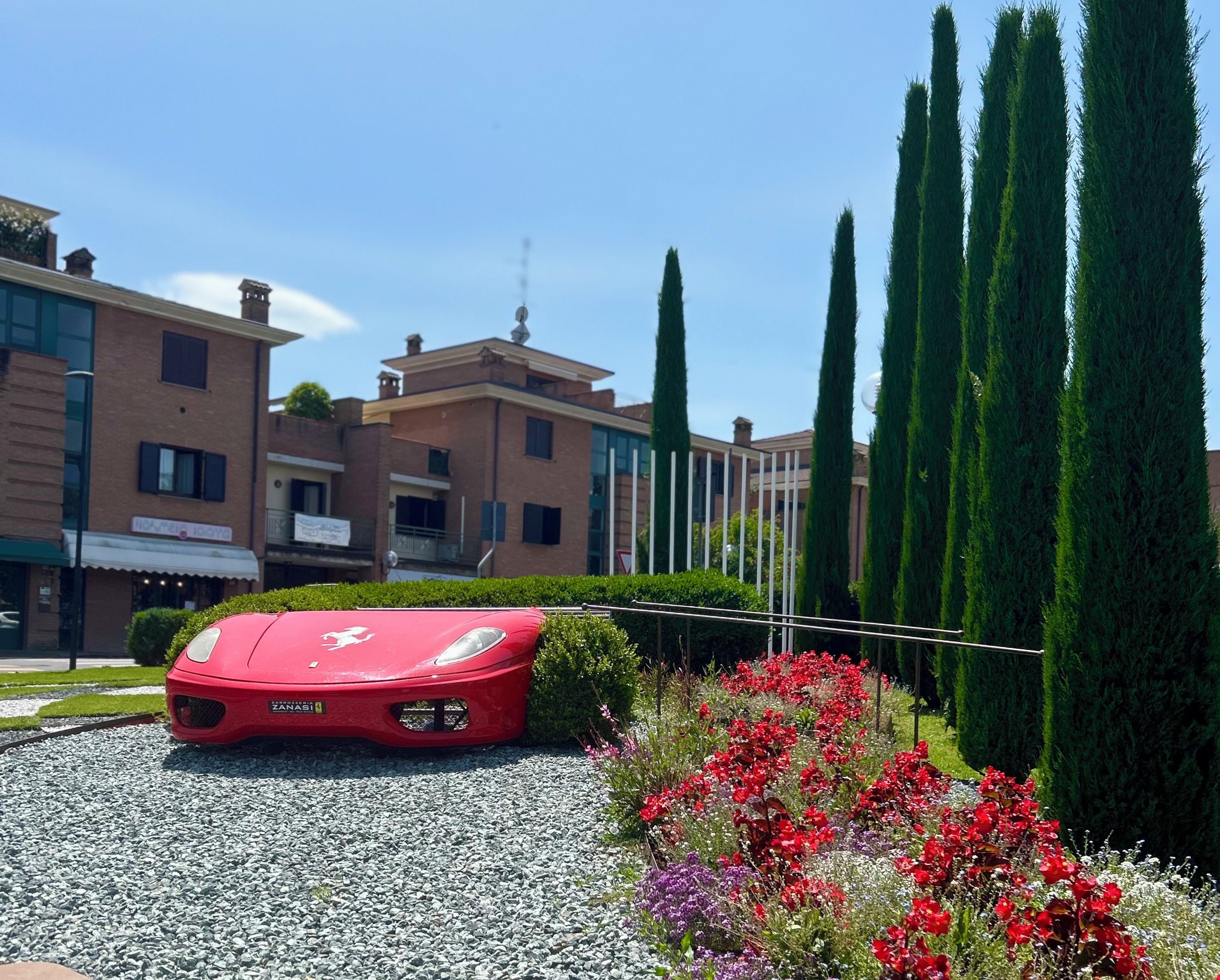 A red sports car parked in a driveway with a gravel surface, surrounded by a garden with red flowers and tall green trees.