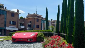A red sports car parked in a driveway with a gravel surface, surrounded by a garden with red flowers and tall green trees.