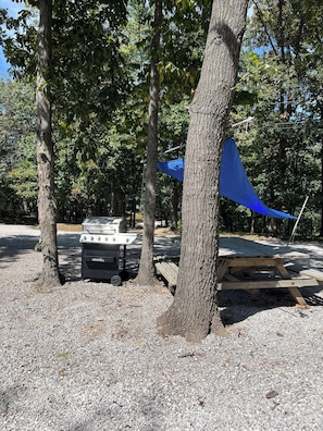 A camping area with a grill, a shaded area, and a wooden bench.