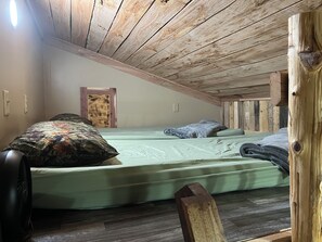 A single bed with a green mattress and pillows, wooden ceiling beams, a small wooden door, and a wall-mounted light fixture.
