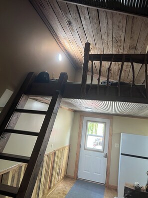 A loft bedroom with a wooden ceiling, a ladder, a window, and a door.
