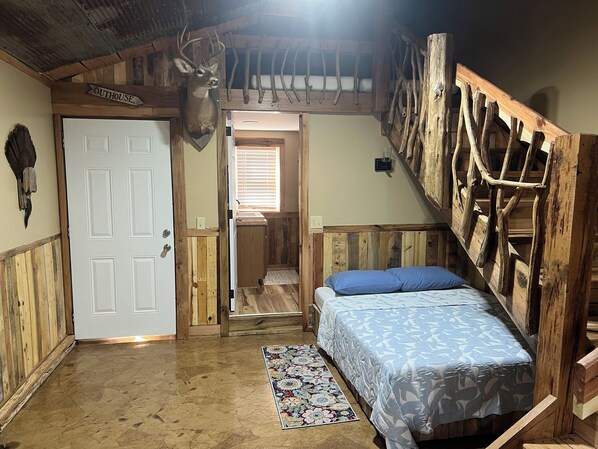 Une pièce rustique avec un lit, une échelle en bois, une tête de cerf accrochée au mur et une porte portant l’inscription « Toilettes extérieures ».