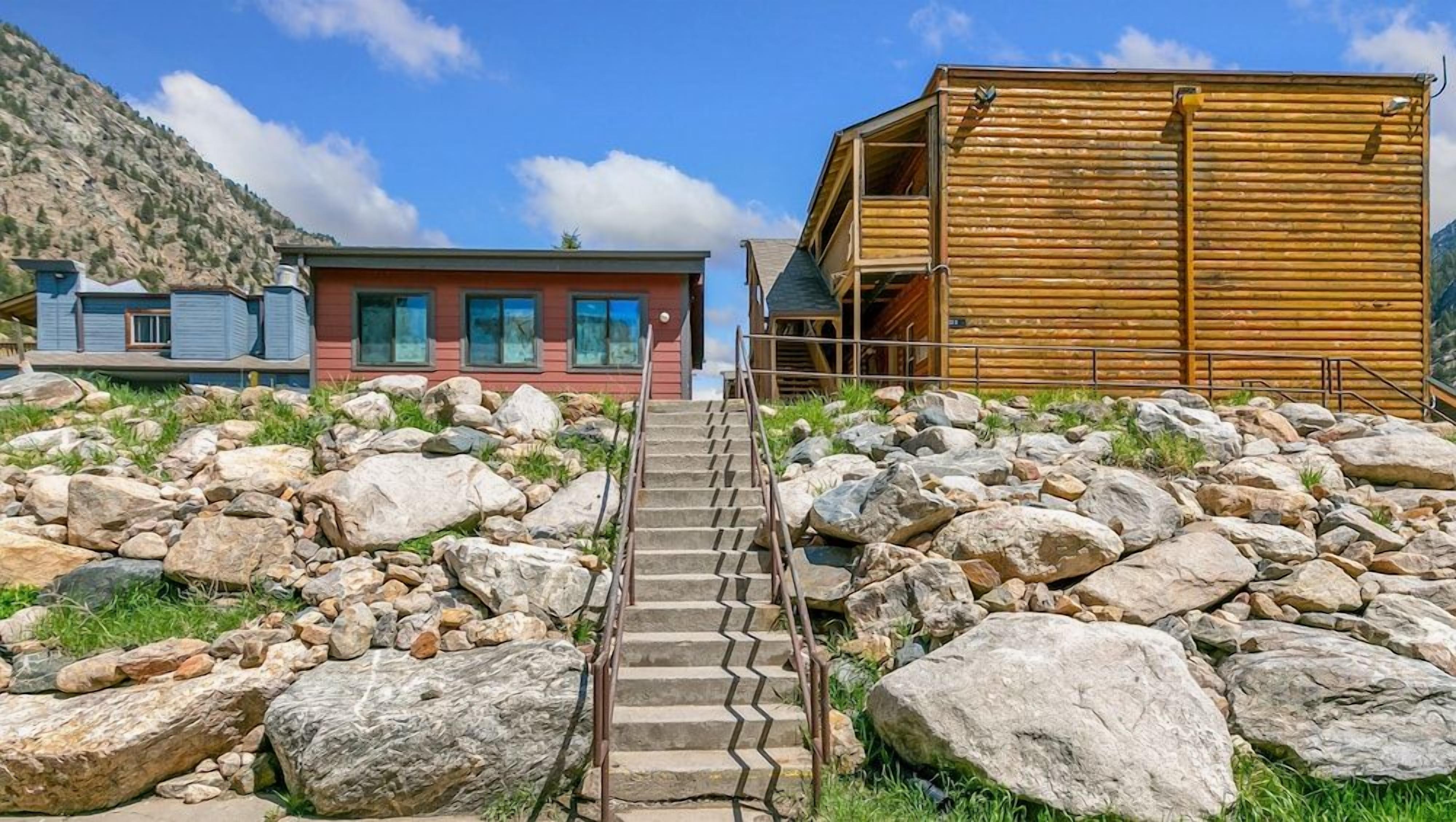 A building with a staircase leading to a wooden structure, surrounded by large rocks and a mountain in the background.