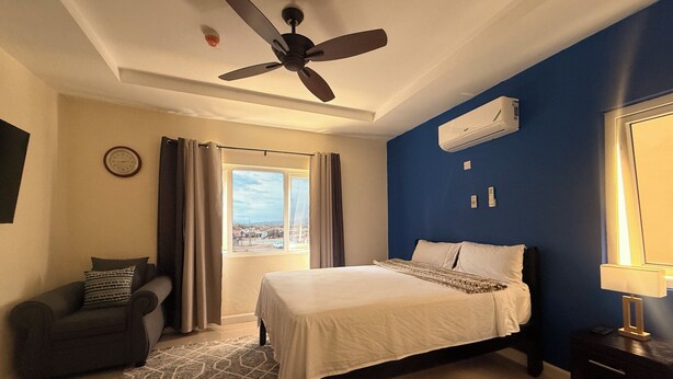 Habitación de hotel con una cama, ventilador de techo, unidad de aire acondicionado, ventana con cortinas, un reloj y una alfombra estampada.