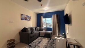 Una sala de estar con un sofá, una mesita, un ventilador de techo, una ventana con cortinas y un cuadro en la pared.