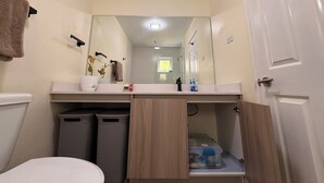 Un baño con inodoro, lavamanos con espejo, toallero y cubo de basura.