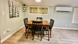 A dining area with a wooden table and chairs, a hanging light fixture, a wall-mounted air conditioner, and a decorative window with patterned curtains. - Cozy Summerville Cottage, Sleeps 6, Pet Friendly, Fully Fenced Back Yard (Rochester)