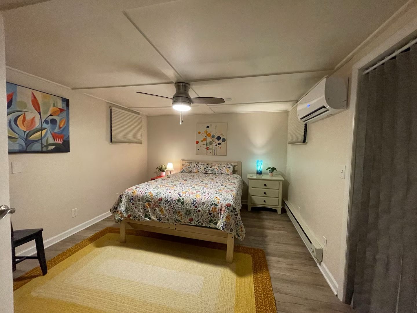 Quarto com cama, ventoinha no teto, mesa de cabeceira, abajur e um quadro na parede.