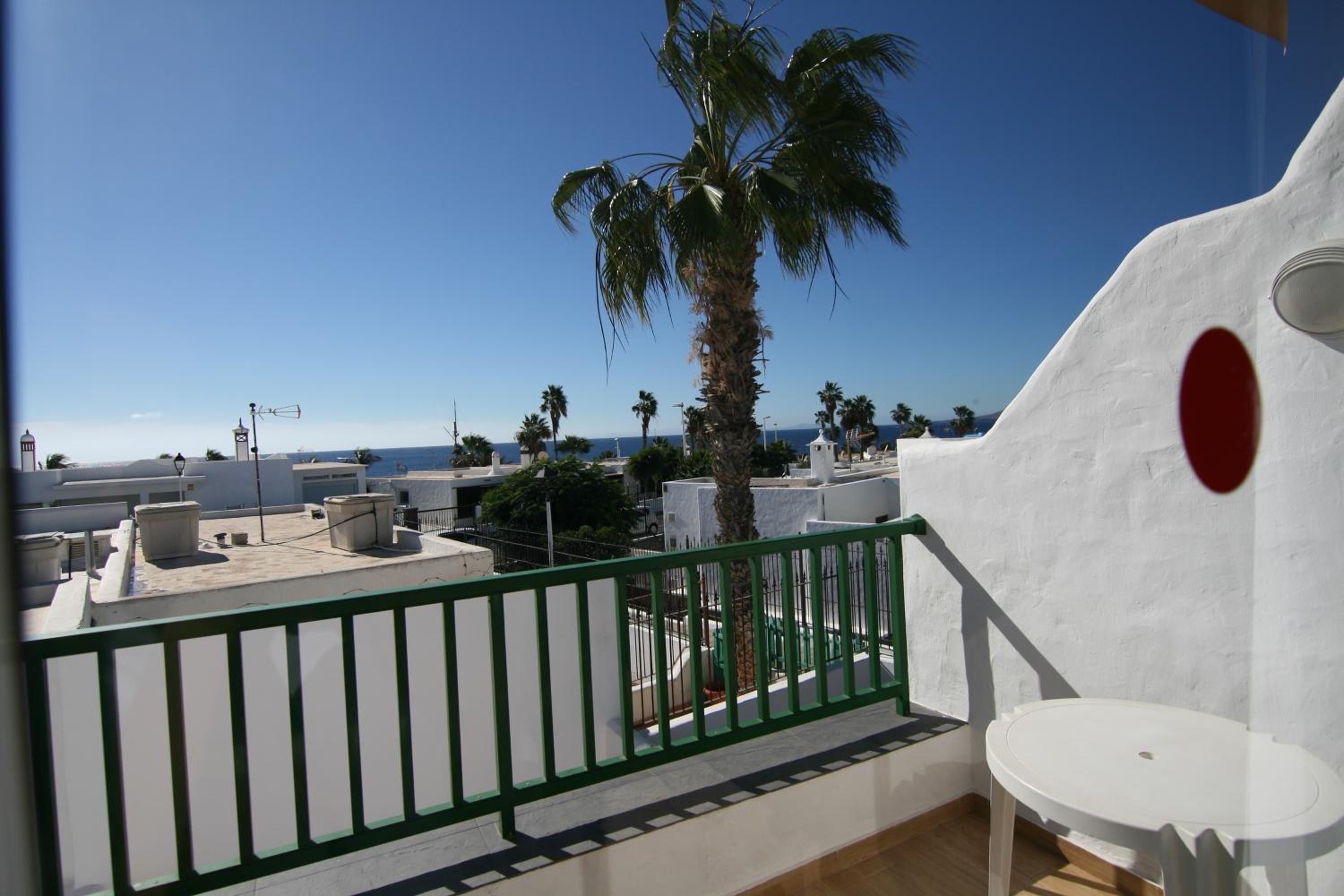 Een balkon met uitzicht op zee, palmbomen en een wit gebouw met een rode cirkel.