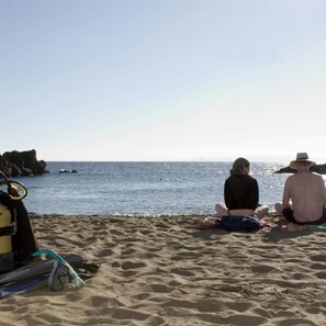 Ein sandiger Strand mit zwei Personen, die zum Meer sitzen, einer Taucherbrille und einer Tasche im Sand.