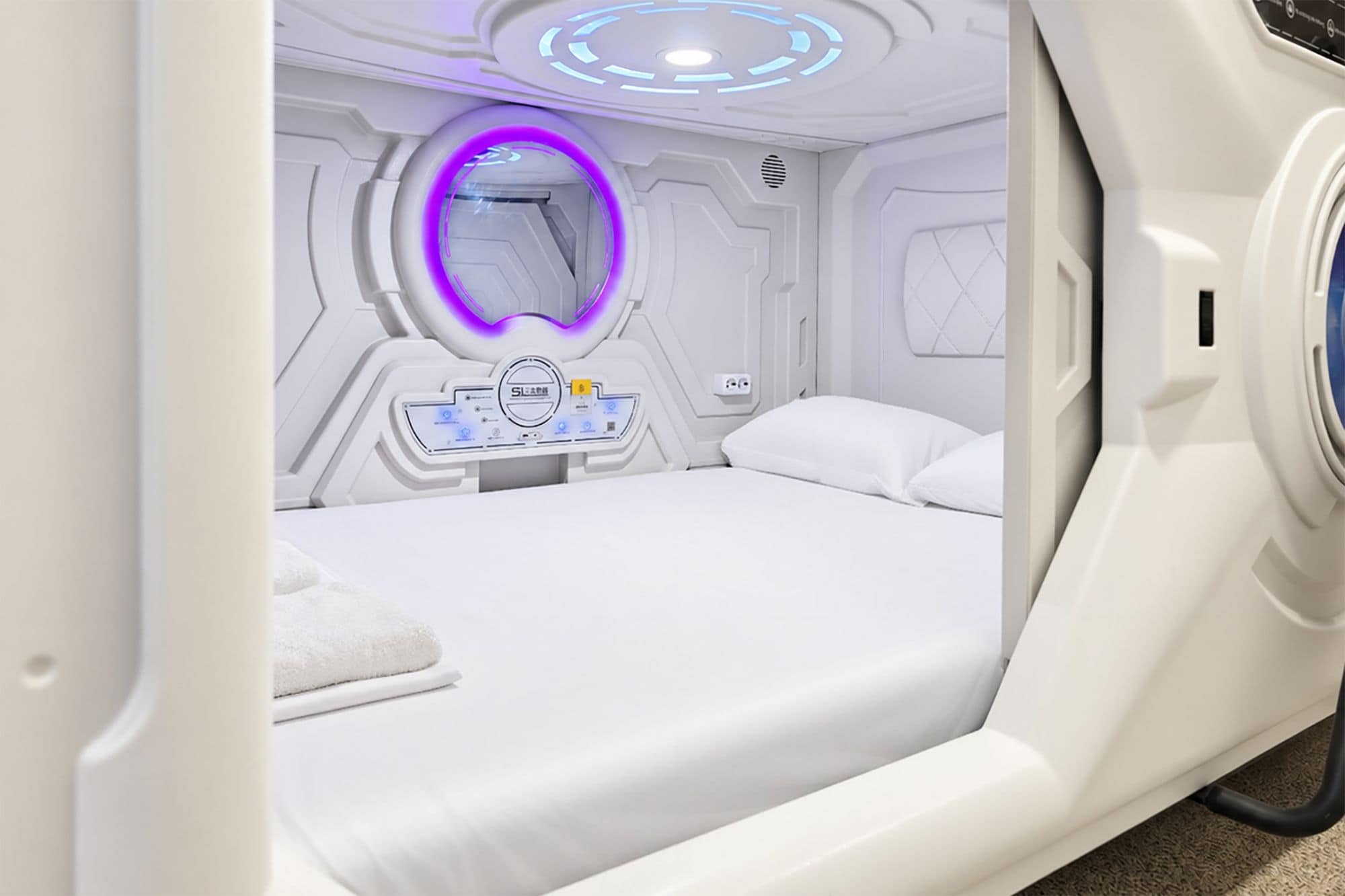 Una camera moderna, bianca e dalla forma a capsula, con un letto, un pannello di controllo e una lampada circolare.