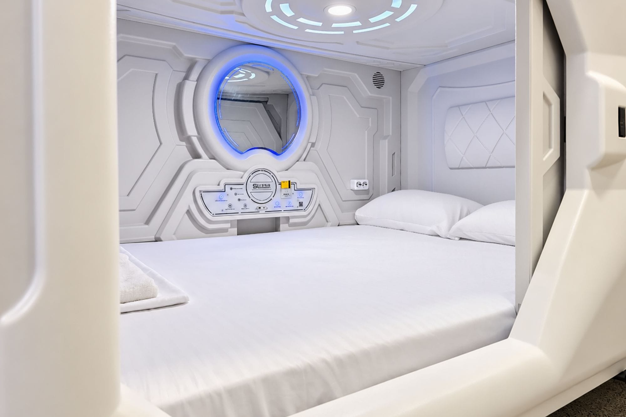Une chambre à coucher moderne, aux tons blancs, dotée d’un panneau de contrôle, d’un luminaire circulaire et d’un lit.
