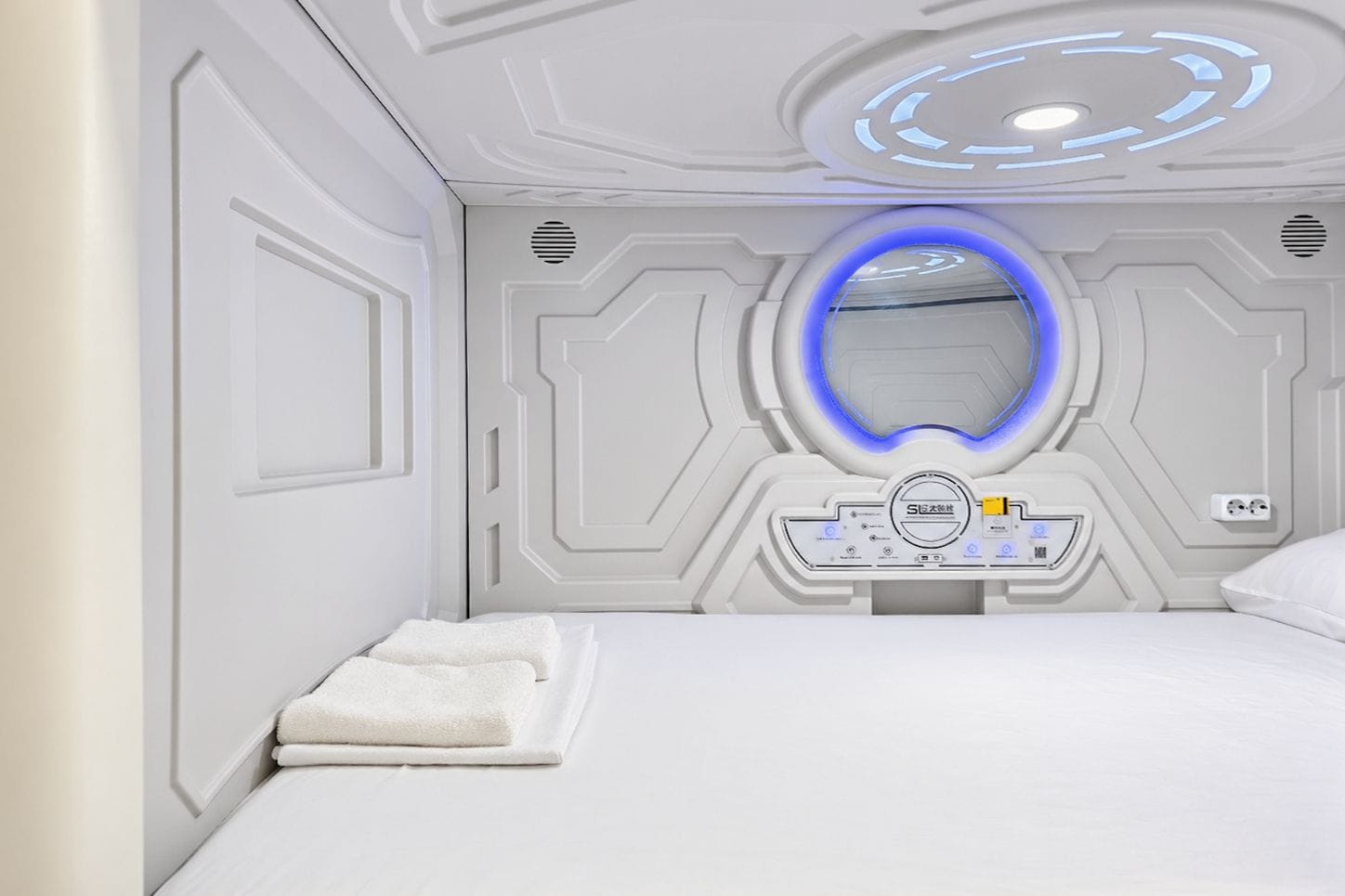 A modern hotel room with a bed, a control panel, and a circular light fixture.